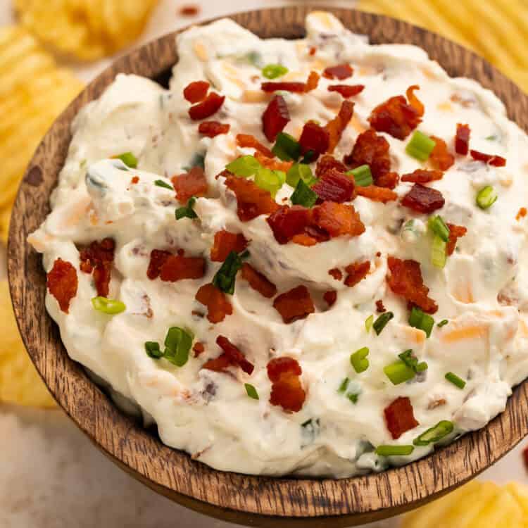 Wooden bowl of loaded baked potato dip surrounded by ruffled potato chips.