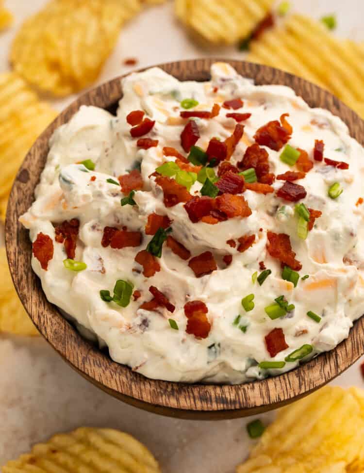 Wooden bowl of loaded baked potato dip surrounded by ruffled potato chips.
