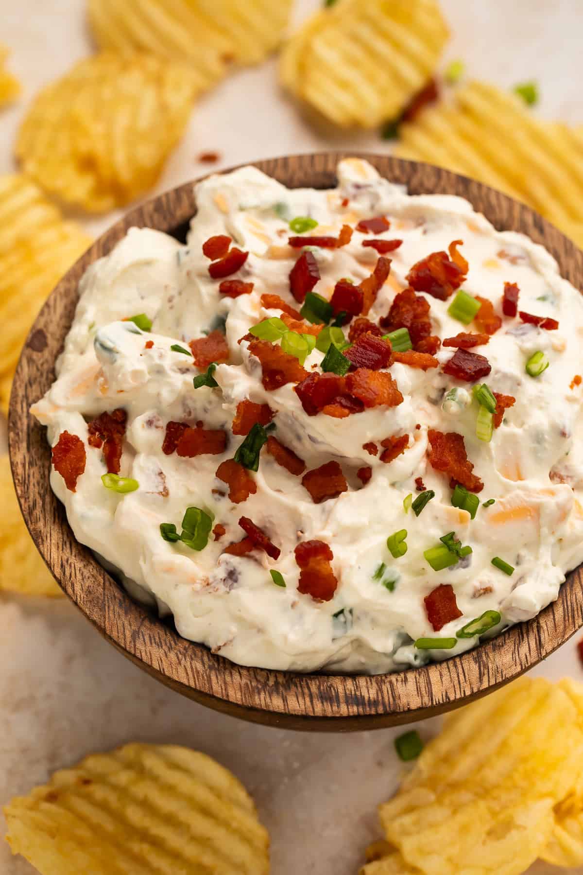 Wooden bowl of loaded baked potato dip surrounded by ruffled potato chips.