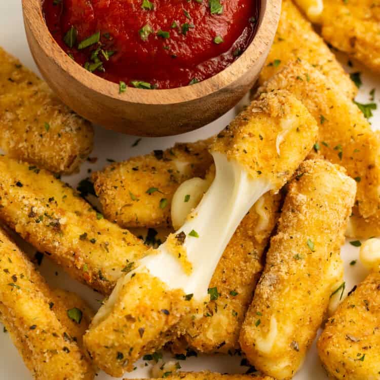 Close up of a pile of air fry mozzarella sticks with a wooden bowl of tomato sauce. One of the mozzarella sticks is broken in half to show the cheese pull.