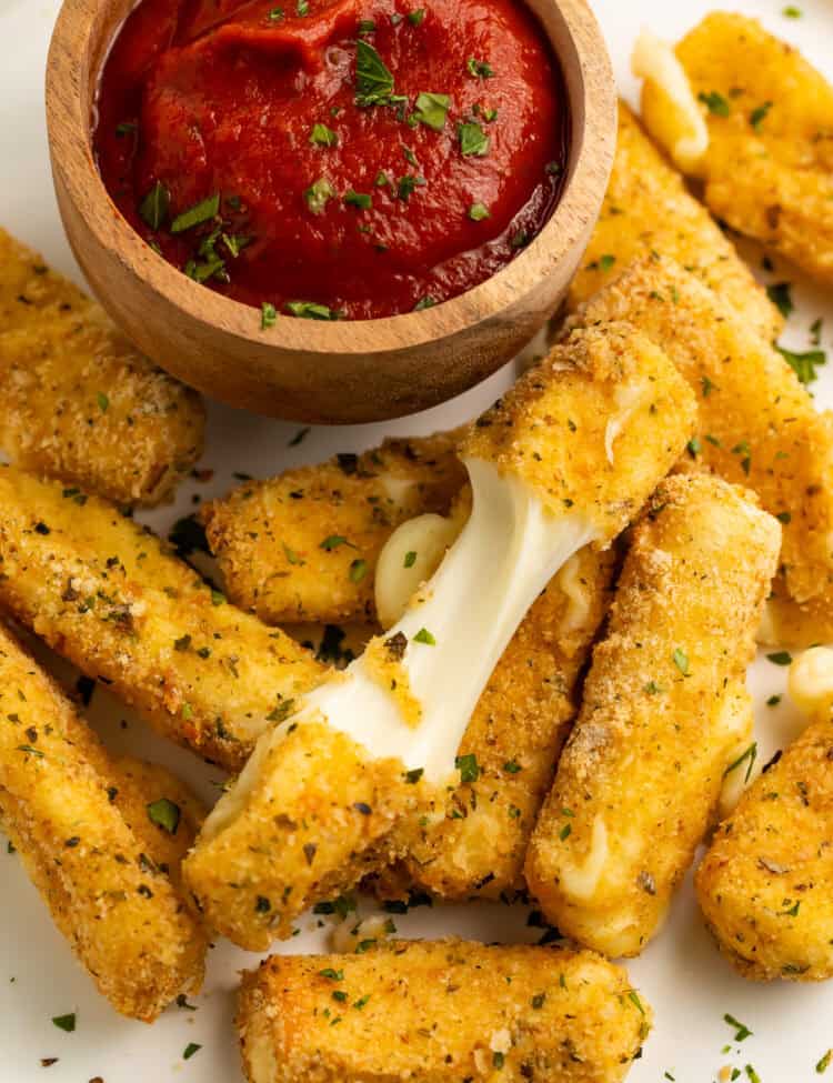 Close up of a pile of air fry mozzarella sticks with a wooden bowl of tomato sauce. One of the mozzarella sticks is broken in half to show the cheese pull.