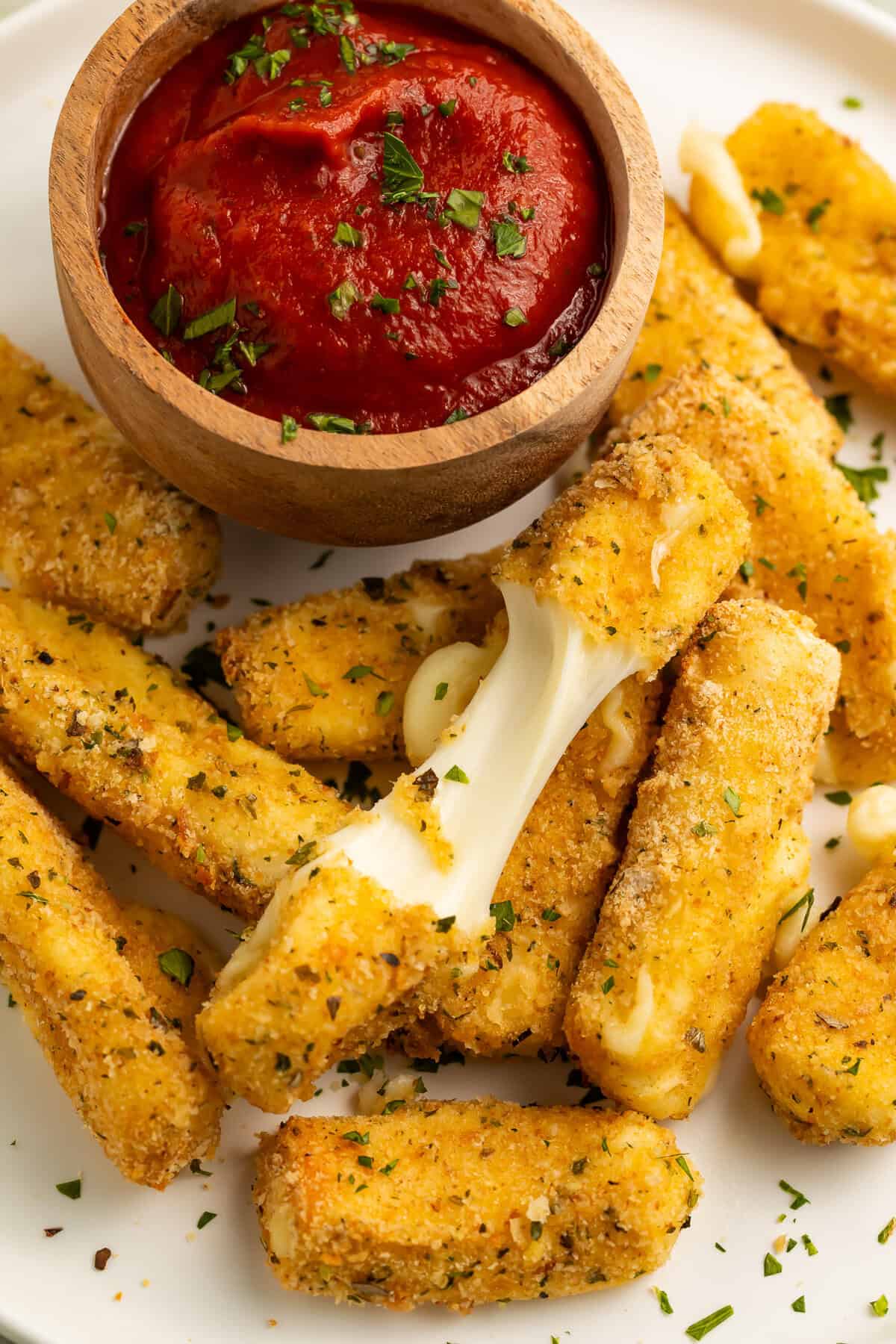 Close up of a pile of air fry mozzarella sticks with a wooden bowl of tomato sauce. One of the mozzarella sticks is broken in half to show the cheese pull.