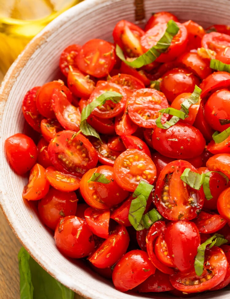 Top-down view of a bowl of rich, bright red tomatoes with fresh green basil in a white bowl.