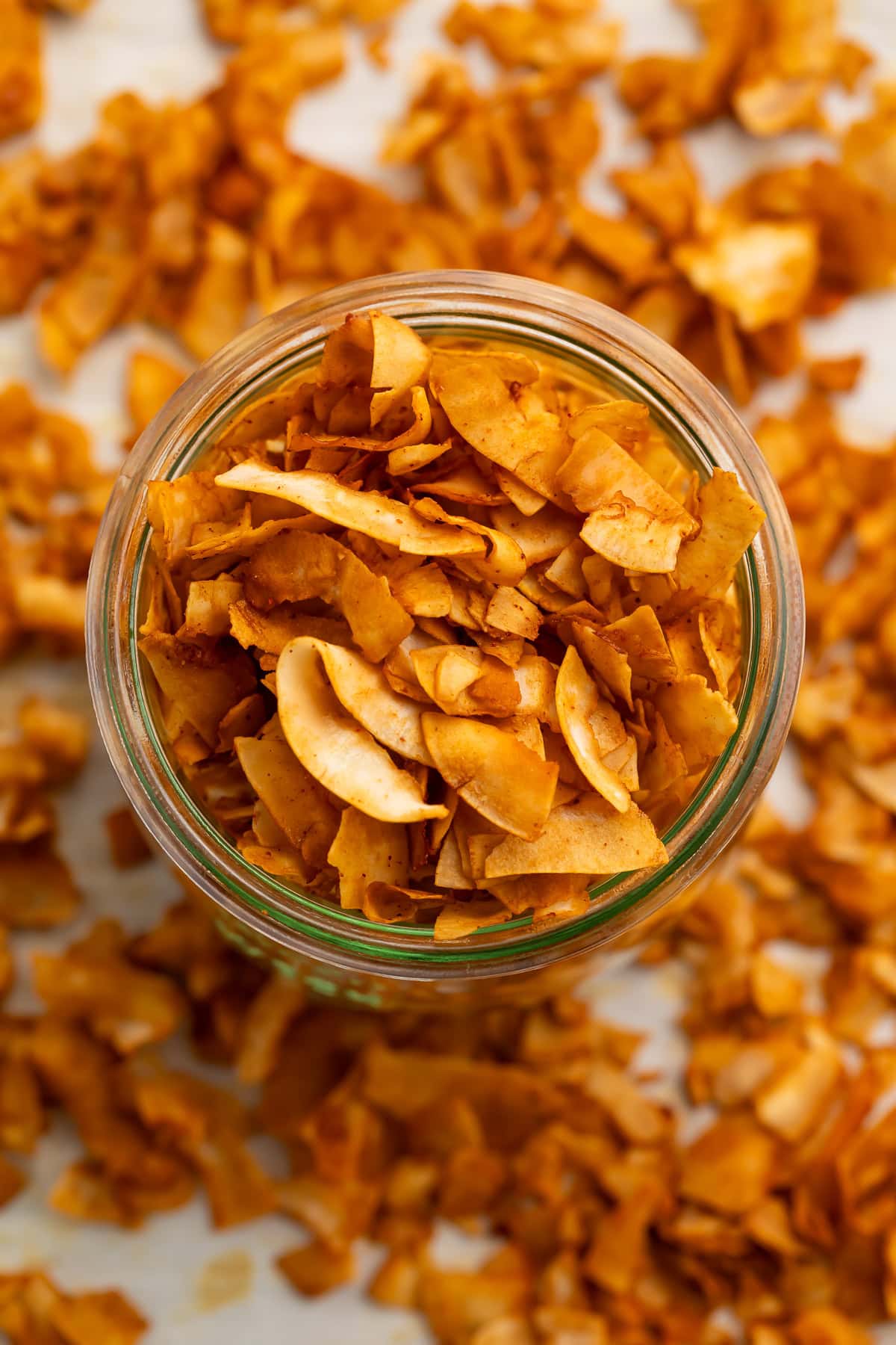 A glass jar of crispy coconut bacon chips on a table surrounded by coconut bacon.