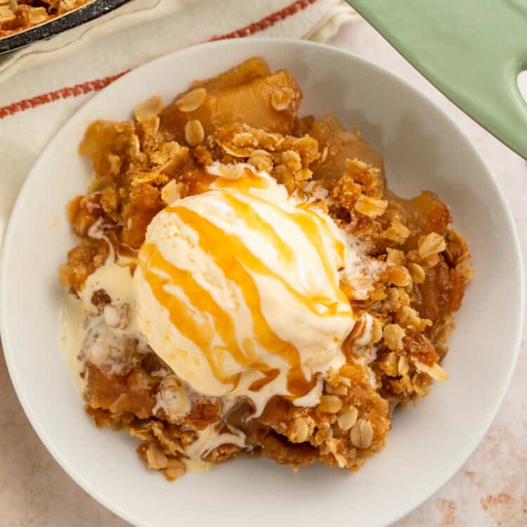 White bowl of apple crisp topped with a scoop of vanilla ice cream and a caramel drizzle.