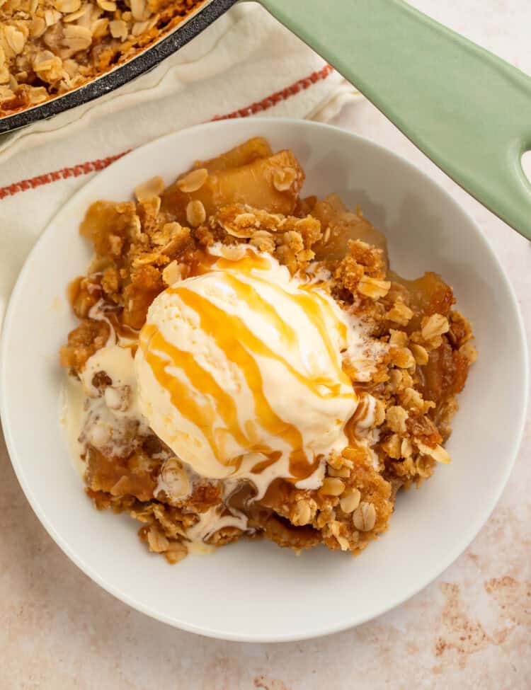 White bowl of apple crisp topped with a scoop of vanilla ice cream and a caramel drizzle.