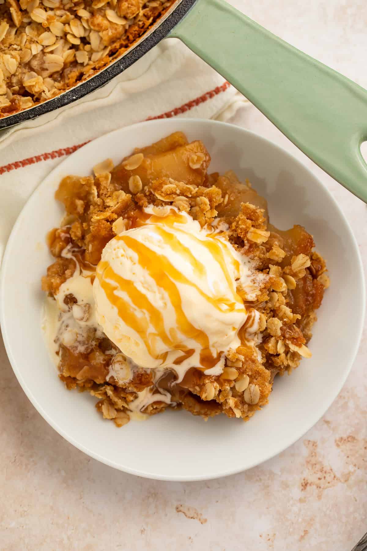 White bowl of apple crisp topped with a scoop of vanilla ice cream and a caramel drizzle.