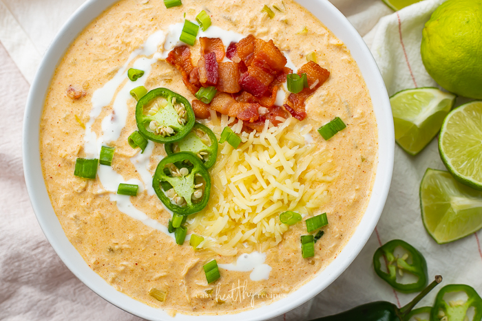 A bowl full of keto chicken white chili topped with pieces of bacon and jalapeno slices next to lime wedges