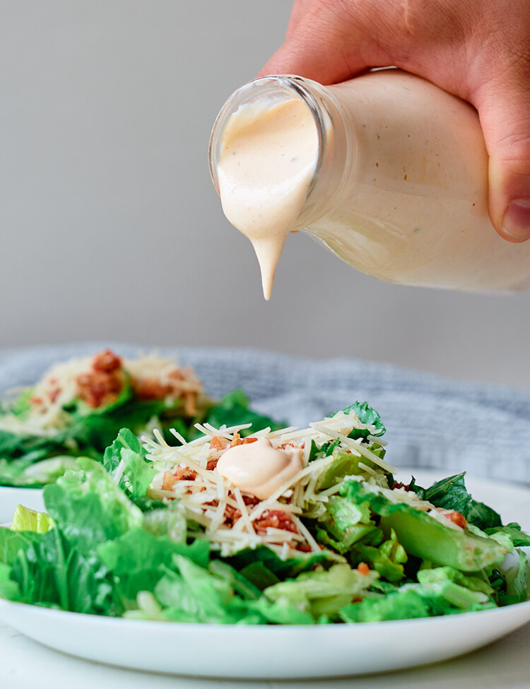 A hand pouring buffalo ranch dressing from a jar onto a salad