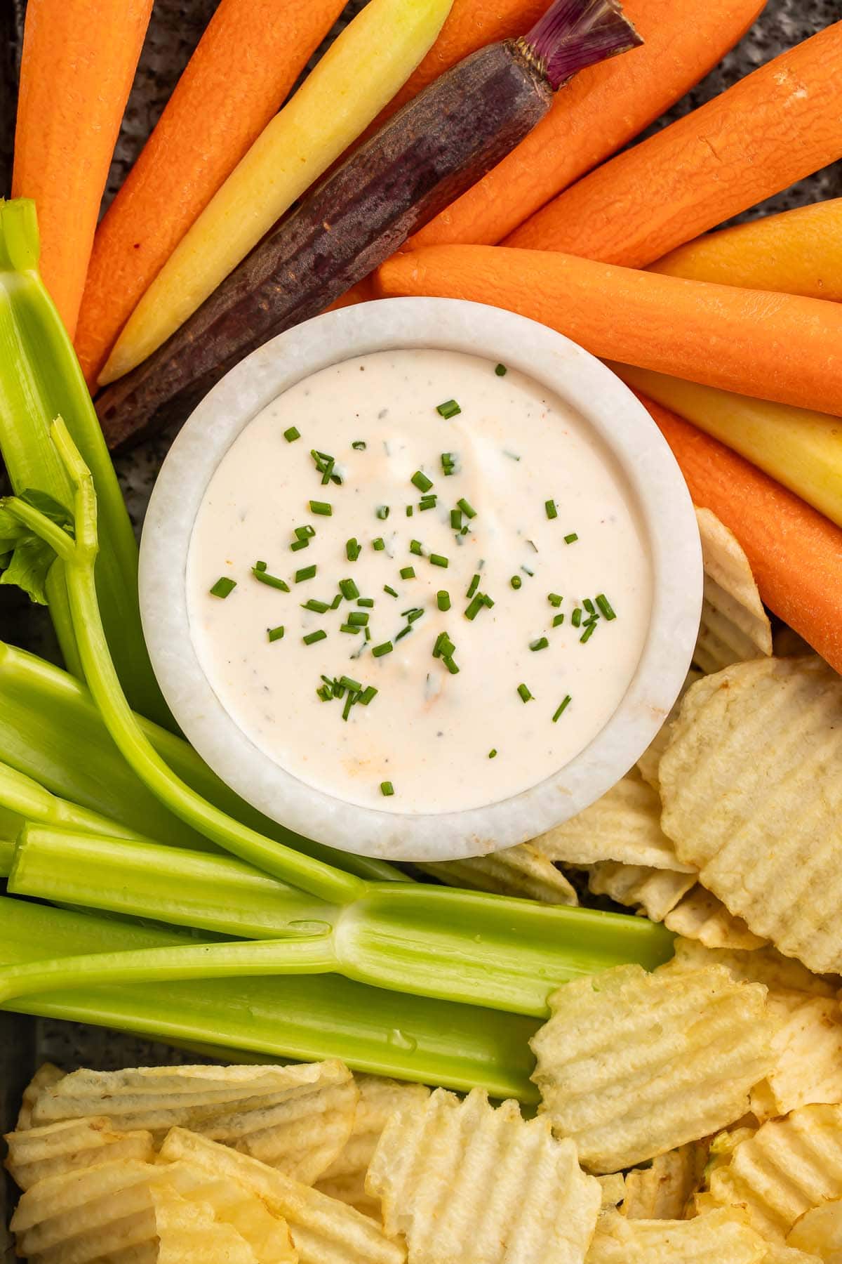 A bowl of buffalo ranch dressing surrounded by carrot sticks, celery sticks, and potato chips on a pan.