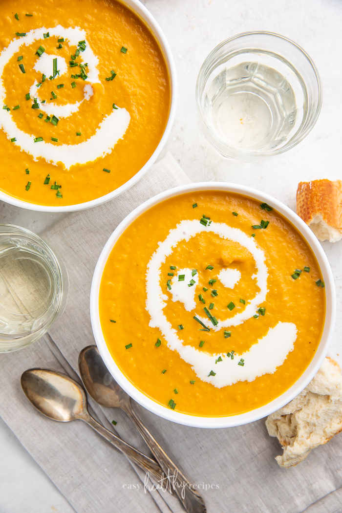 Two bowls of pumpkin bisque surrounded by spoons, glasses, and bread
