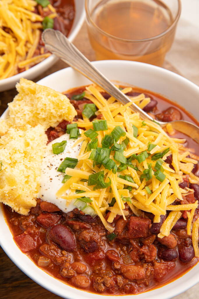 A bowl of bison chili in a white bowl topped with cheese and sour cream with a side of cornbread