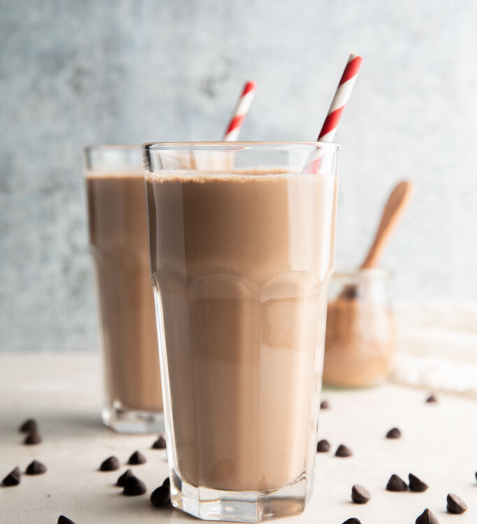 Two glasses of keto chocolate milk surrounded by chocolate chips with red and white striped straws