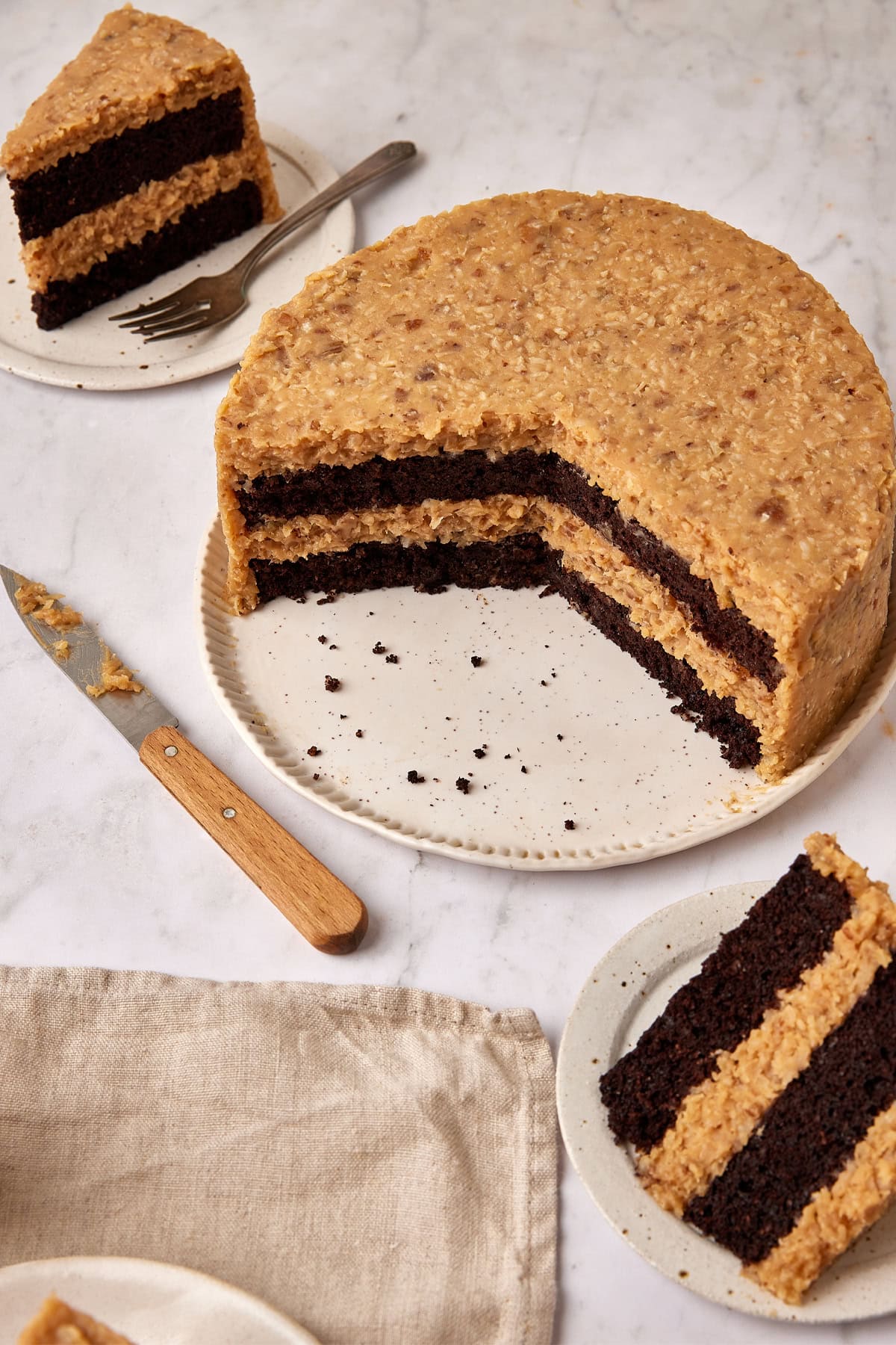 A 2/3 cut German Chocolate Cake with slices on plates, a napkin and a knife surrounding it on the countertop. 