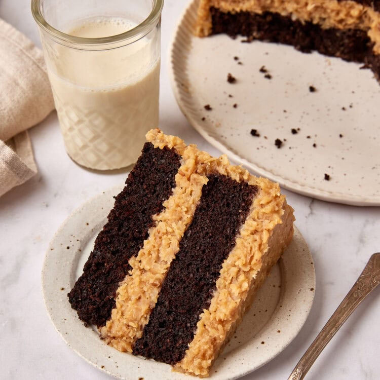 Slice of German Chocolate cake sits on a small white plate net to a fork. A glass of milk is sitting next to it and the cut cake is just visible in the background.