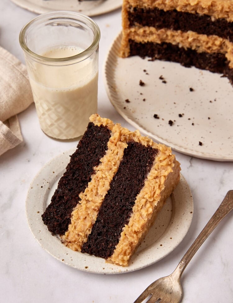 Slice of German Chocolate cake sits on a small white plate net to a fork. A glass of milk is sitting next to it and the cut cake is just visible in the background.