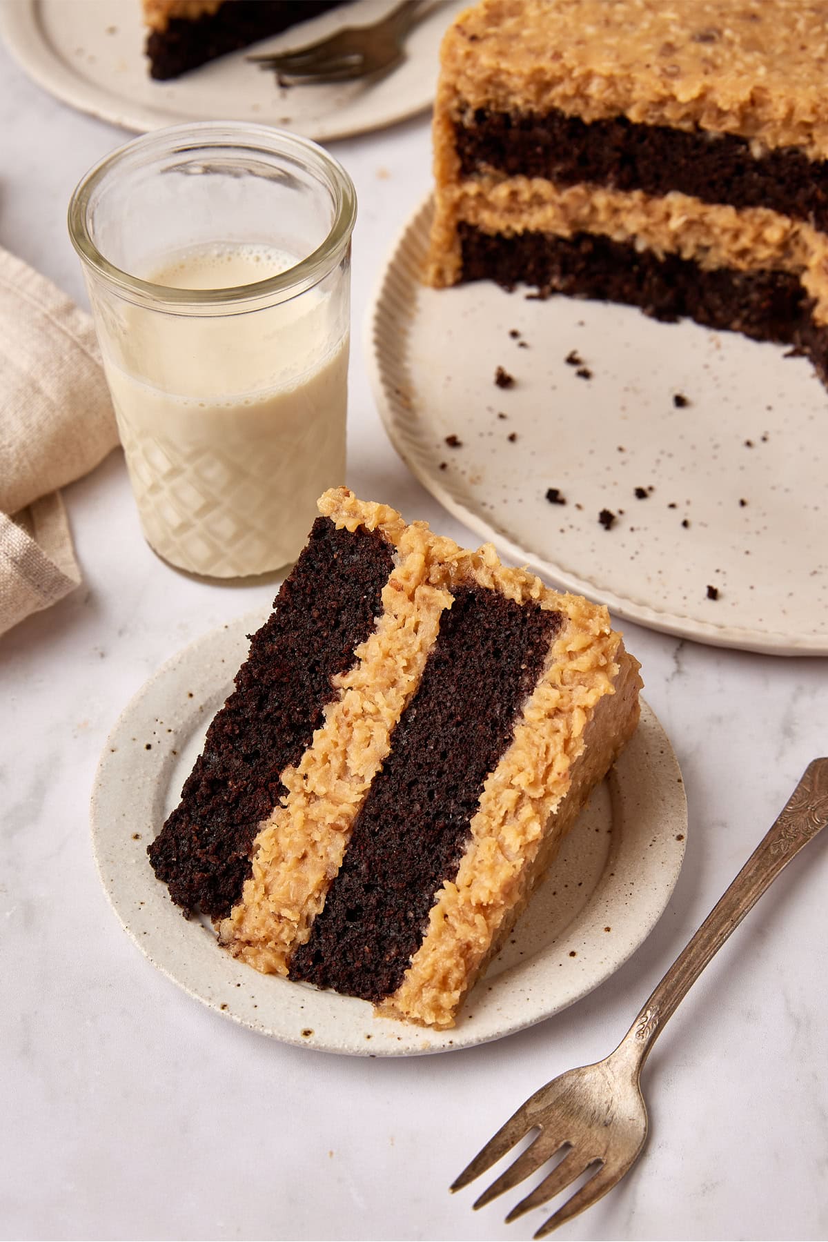 Slice of German Chocolate cake sits on a small white plate net to a fork. A glass of milk is sitting next to it and the cut cake is just visible in the background.