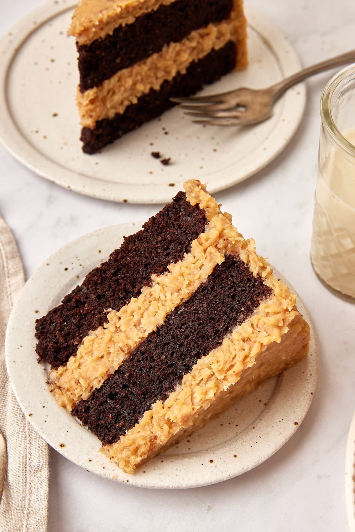 Thick slice of German Chocolate Cake on a white plate, with another slice on a plate with a fork in the background. 