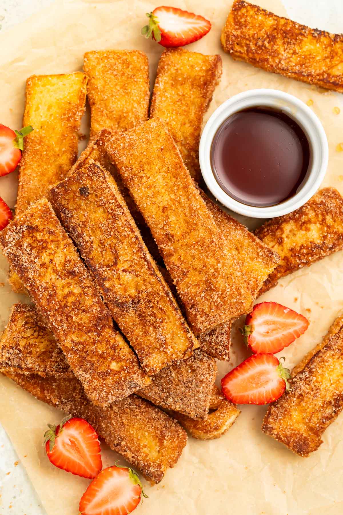 Keto French toast sticks arranged in a pile on a sheet of parchment paper. A ramekin of keto syrup and slices of strawberries sit next to the french toast sticks.