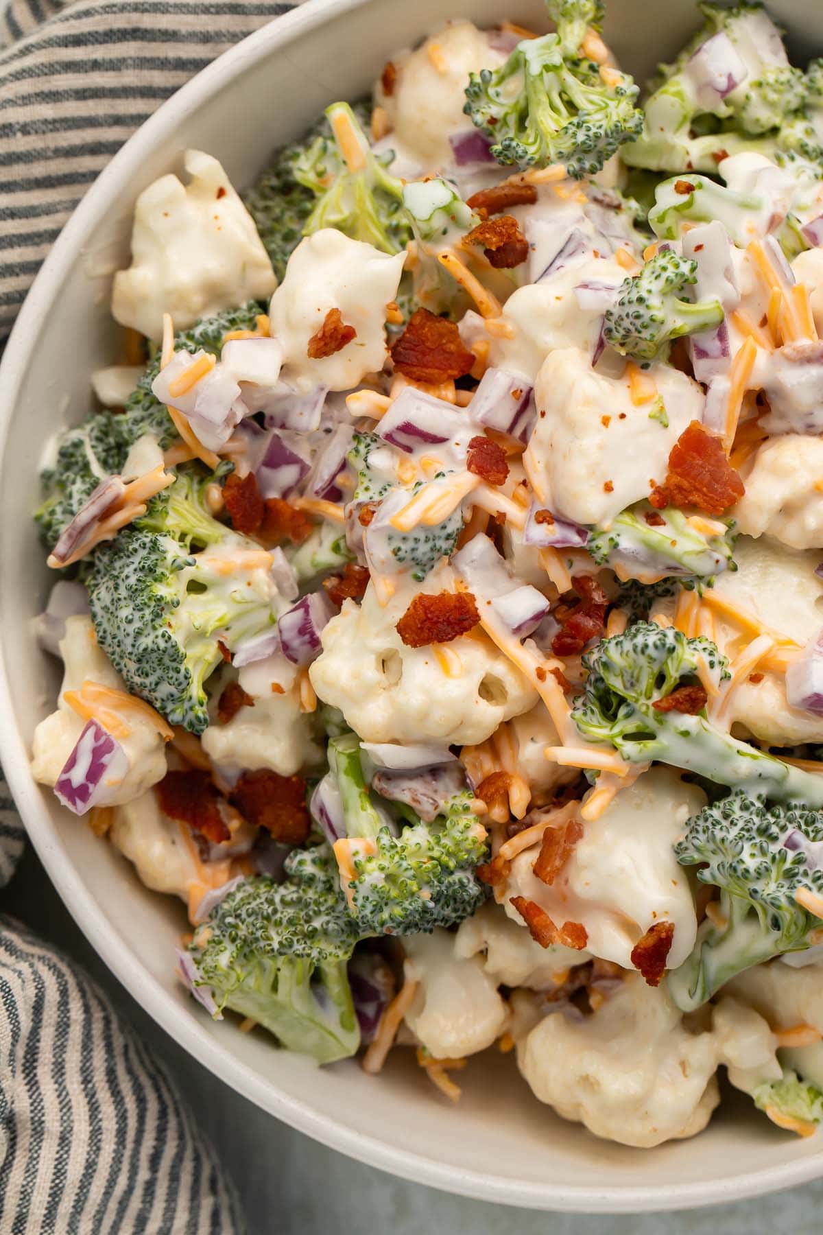 A large white bowl of Amish broccoli salad made with broccoli and cauliflower florets, bacon, cheddar cheese, and mayo.