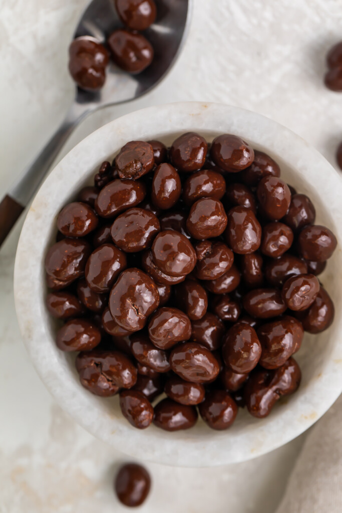 Chocolate covered espresso beans in a small white bowl next to a silver spoon