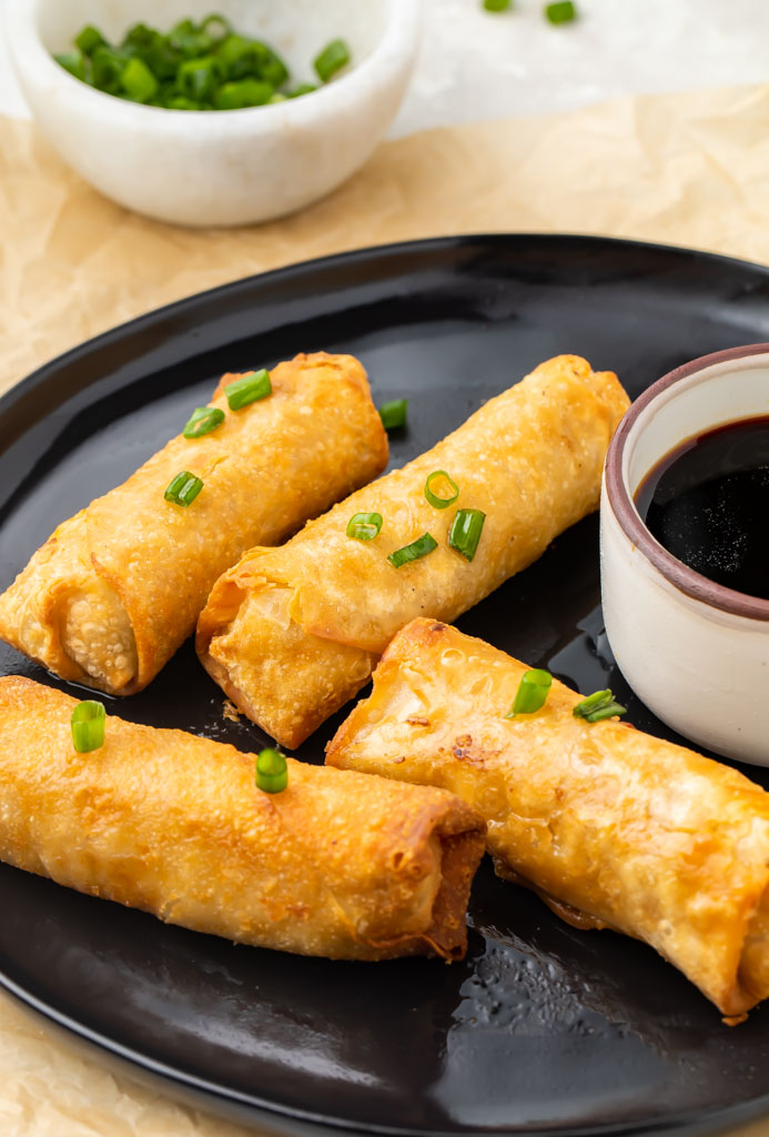4 air fried egg rolls on a black plate with a ramekin of soy sauce for dipping