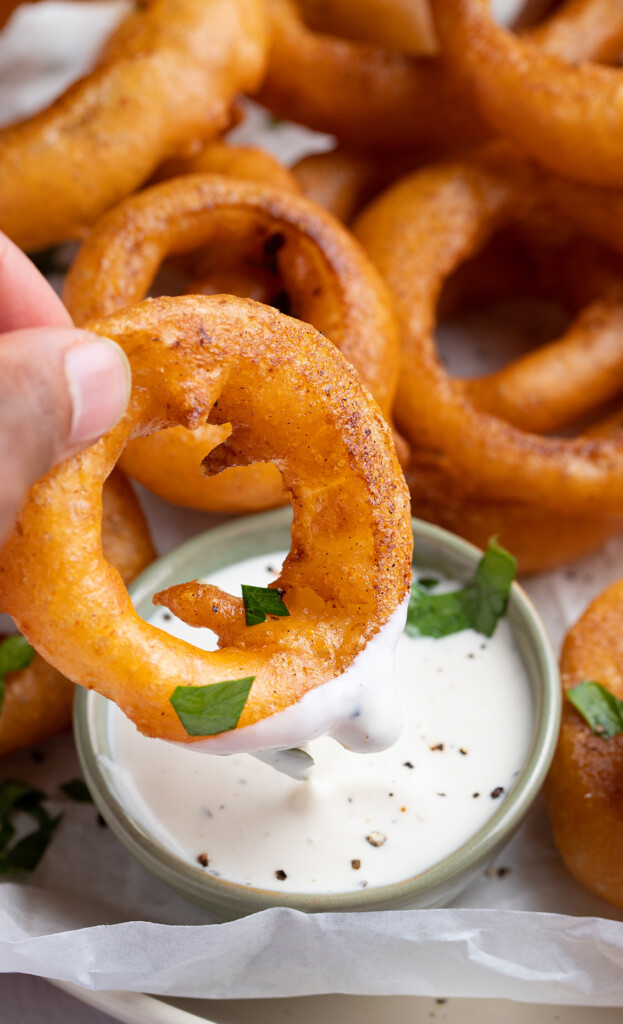 Individual onion ring being dipped into ranch dressing, with more onion rings out of focus in the background