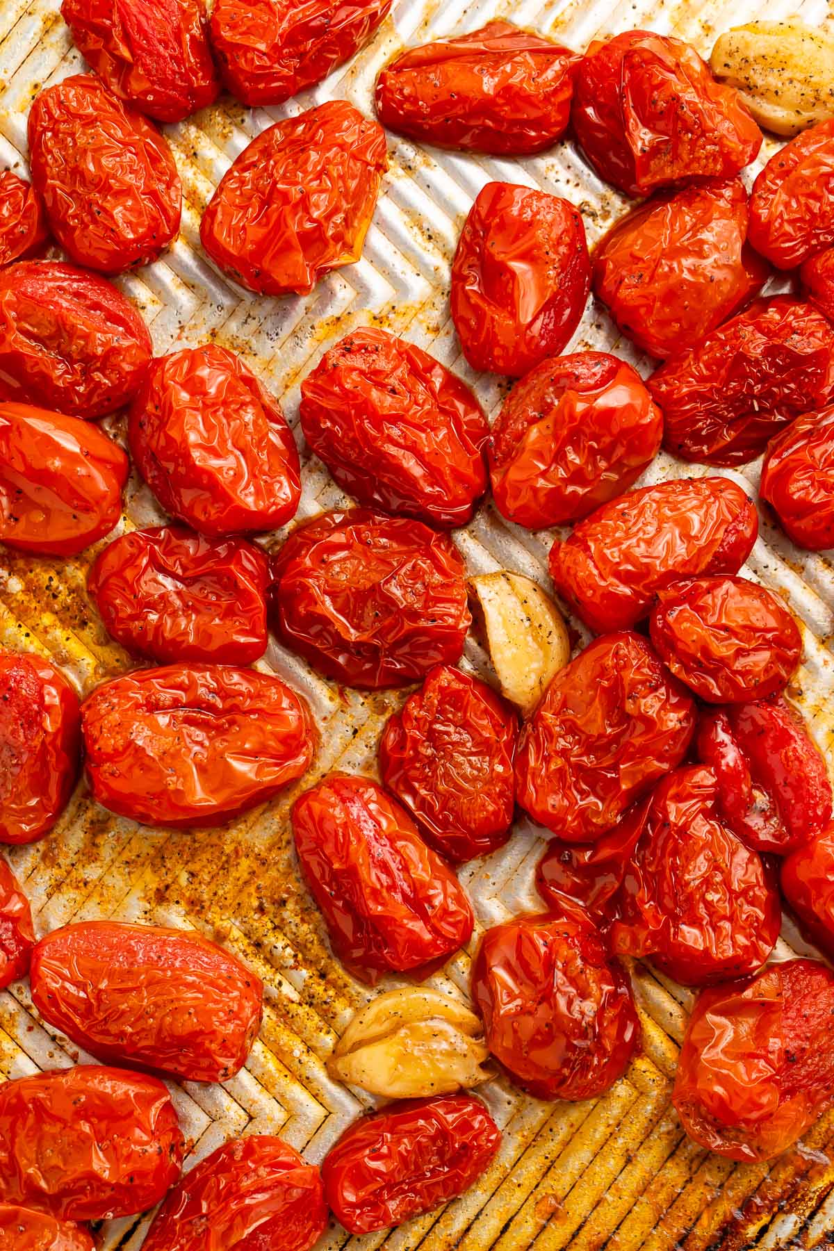 Wrinkled, roasted grape tomatoes and garlic cloves on a silver baking sheet.