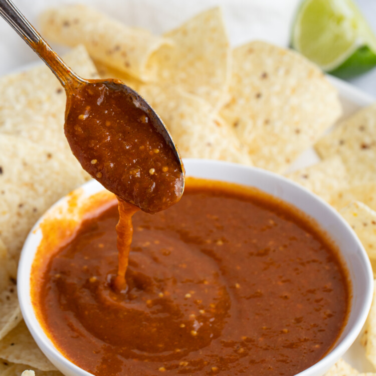 Tomatillo red chili salsa drizzling off a spoon into a white bowl of salsa surrounded by corn tortilla chips
