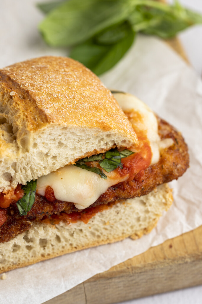 One chicken parm sandwich on a cutting board with parchment paper and spinach in the background