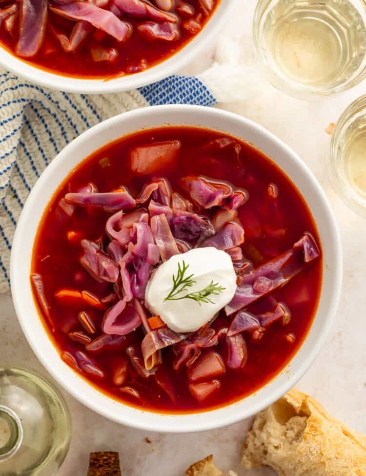 Bowls of red cabbage soup with a dollop of sour cream on top. Cups of wine and torn bread are pictured off to the side.