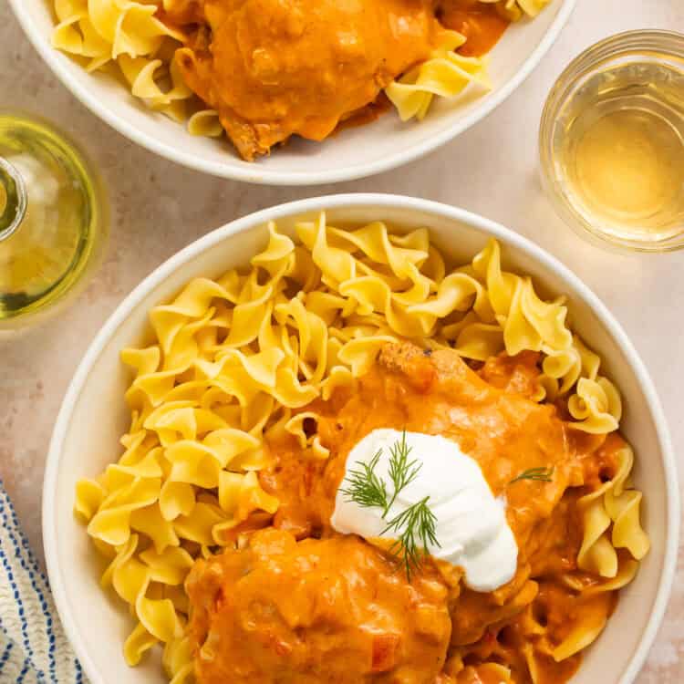 Chicken paprikash chicken thighs with a dollop of sour cream and herbs served over two bowls of egg noodles.