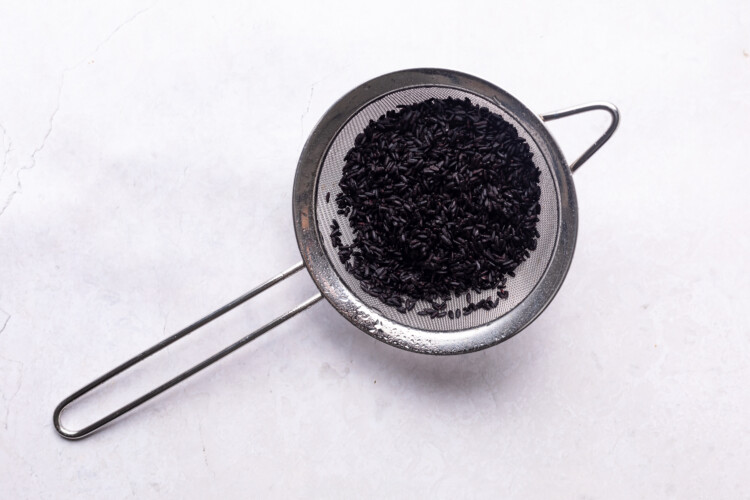 Uncooked black rice in long-handled strainer on white background.