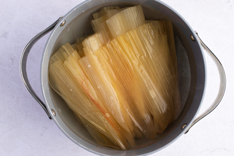 Corn husks in boiling water in large pot on white background.