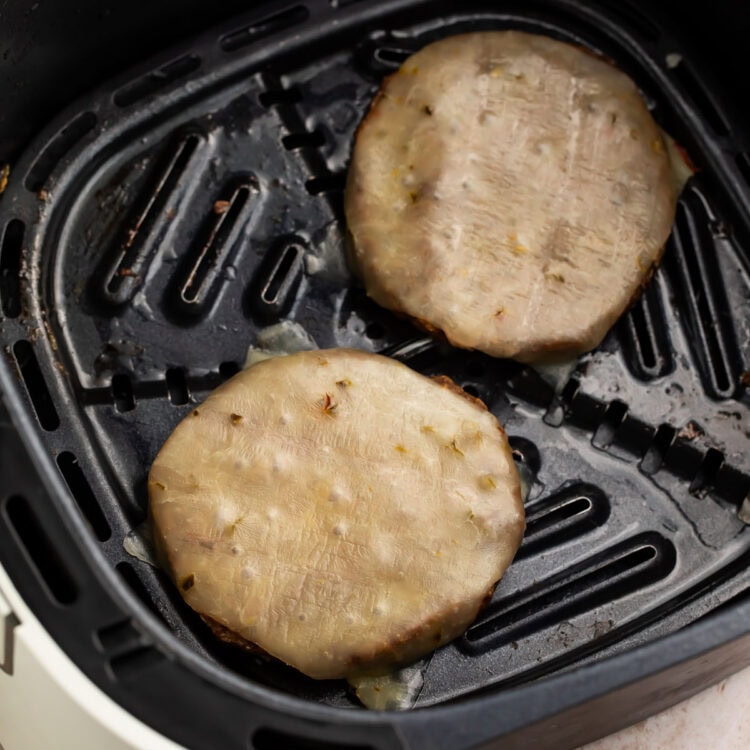 Two frozen burger patties topped with cheese in an air fryer basket.