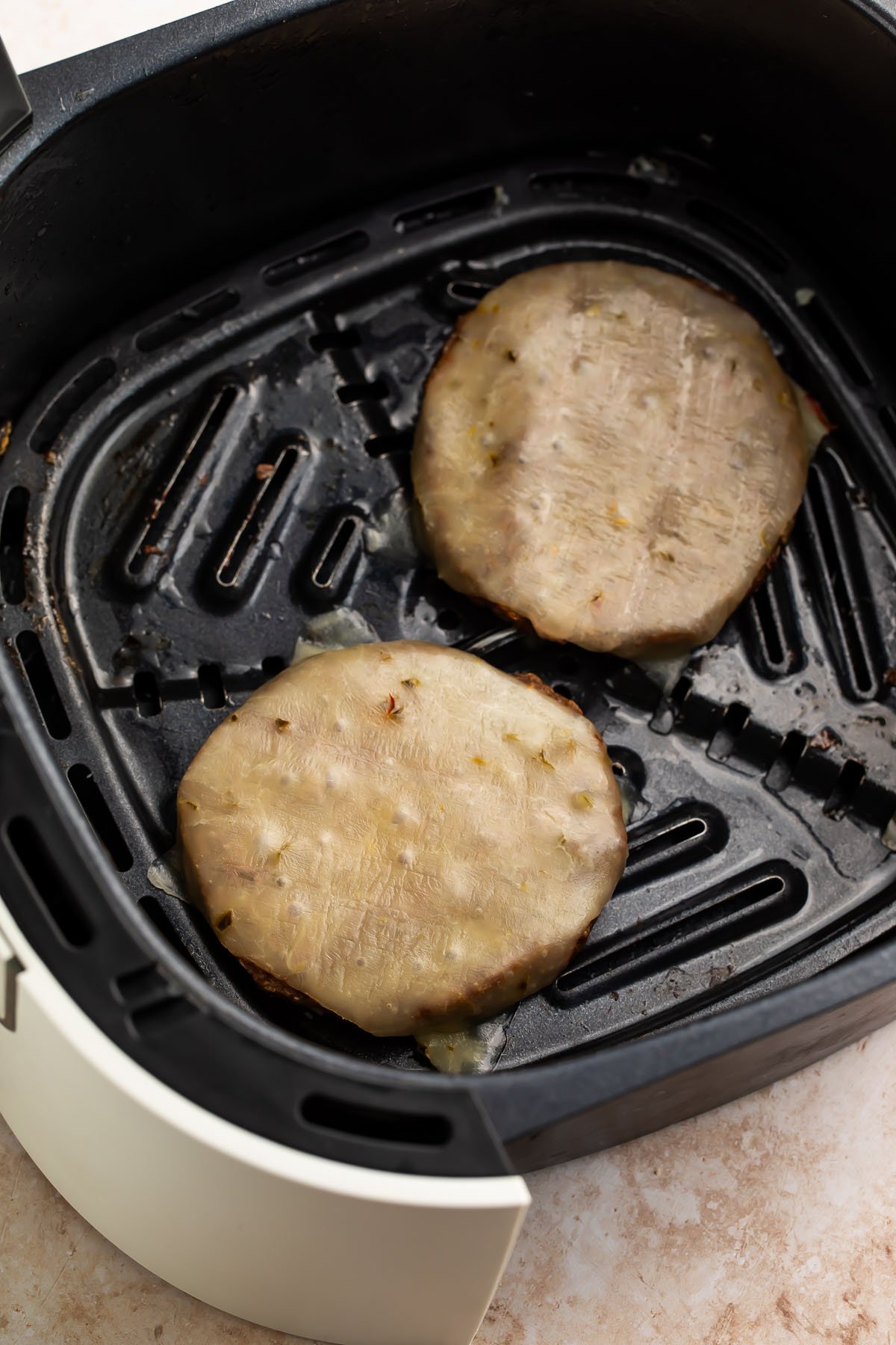 Two frozen burger patties topped with cheese in an air fryer basket.