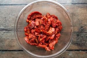 Sliced flank steak in large glass mixing bowl on wooden background.