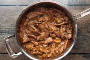 Marinated flank steak in large silver saucepan sitting on a wooden tabletop.