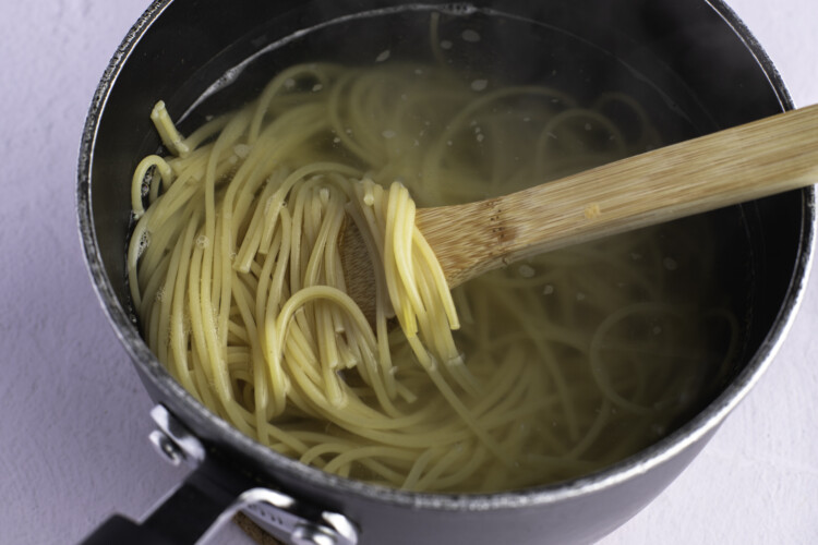 Cooked spaghetti noodles in a large silver pot, with a spoonful of noodles being held up with a wooden spoon.