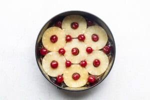 Pineapple rings and maraschino cherries in bottom of cake pan on white table.