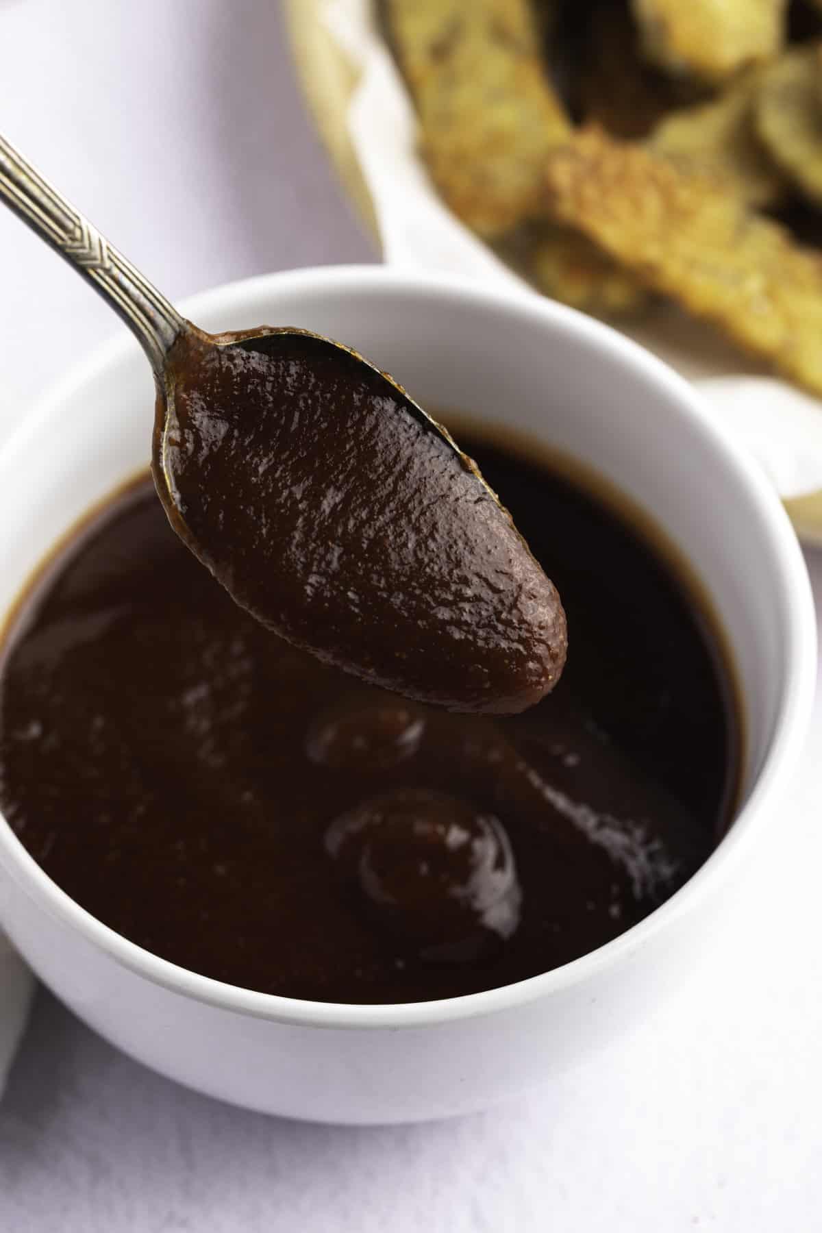 Vegan BBQ sauce in a small white bowl, with a spoon lifting a spoonful of sauce out of the bowl.