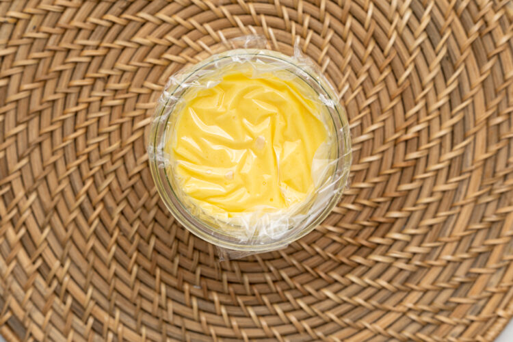 Overhead view of keto lemon curd in a glass jar covered with plastic wrap.