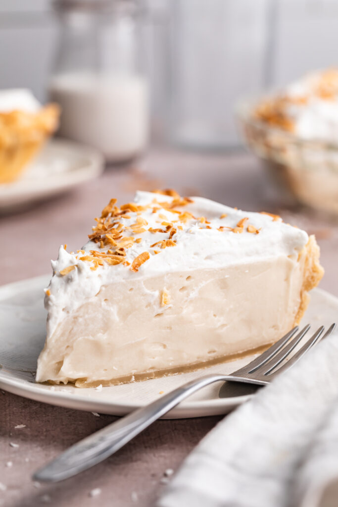 A wedge sliced of vegan coconut cream pie topped with coconut whipped cream and coconut shavings on a neutral plate with a fork.