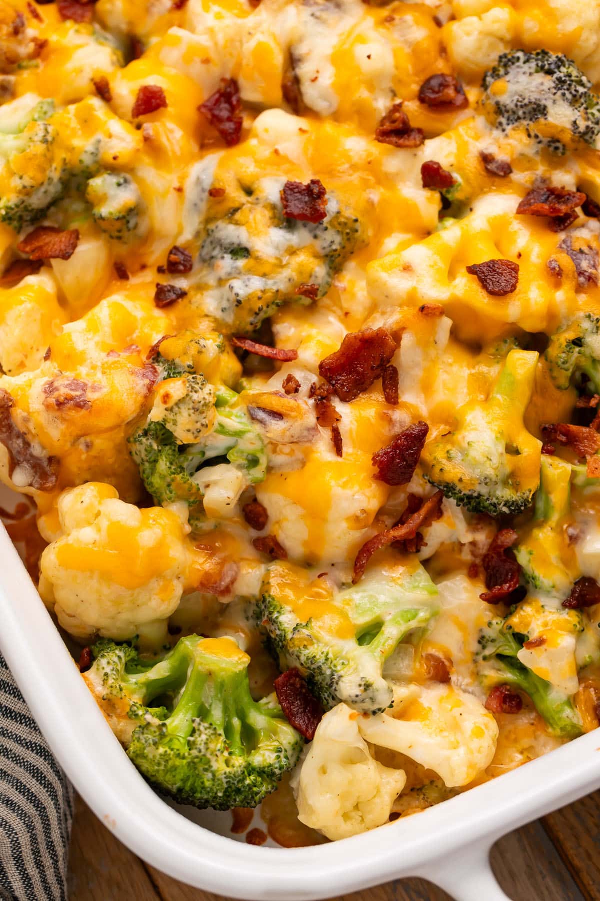 An angled casserole dish, photographed from the corner, holding a keto broccoli cauliflower casserole topped with cheese and bacon pieces.