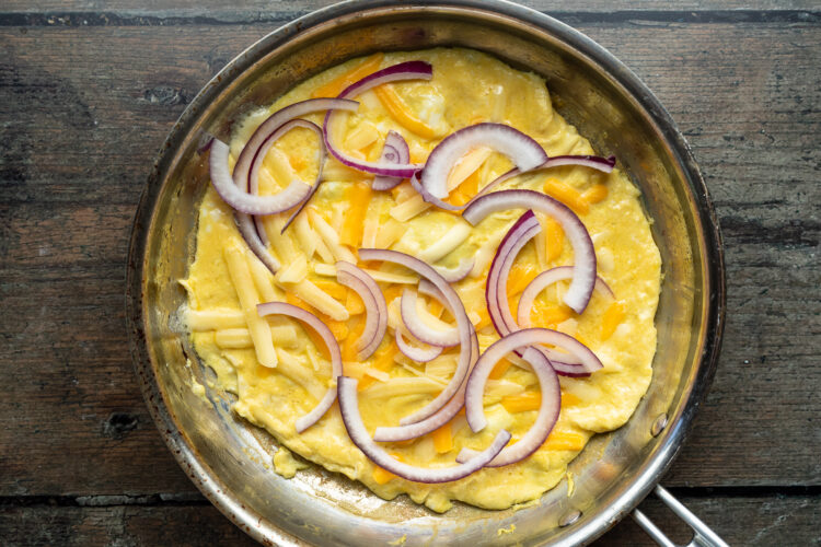 Overhead view of shredded cheddar and sliced red onions on top of an omelette in a skillet.