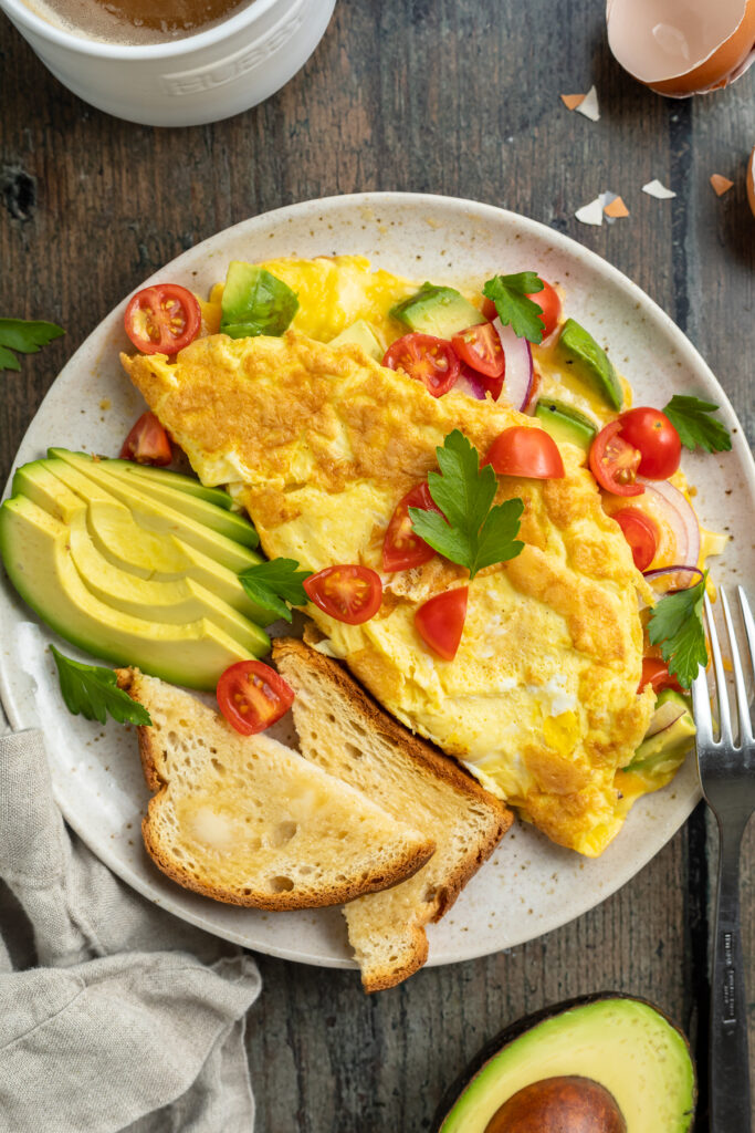 Overhead view of an omelette on a plate with toast, cherry tomatoes, red onions, slices of avocado, and fresh herbs.