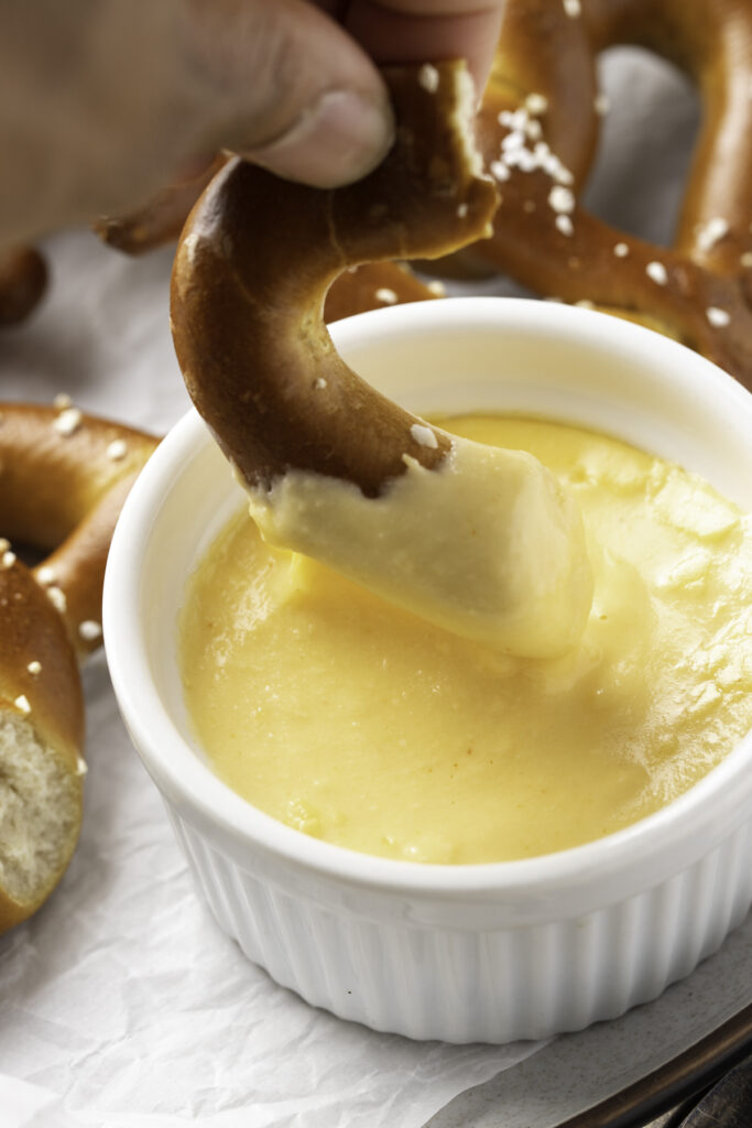 A piece of a salted, twisted, air fryer frozen pretzel being dipped into a small ramekin of cheese sauce.