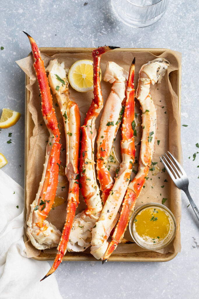 Overhead view of grilled crab legs lined up on a baking sheet lined with parchment paper, next to a small bowl of butter sauce.