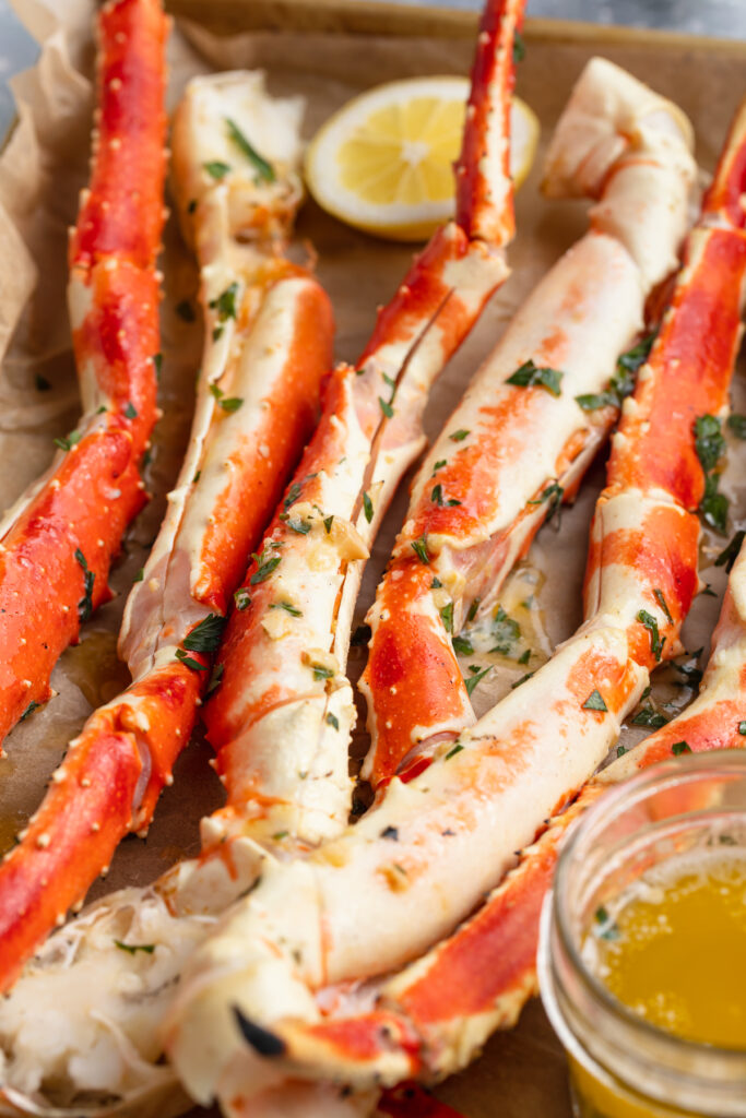 Grilled crab legs, lined up on a baking sheet lined with parchment paper, next to a bowl of garlic butter sauce.