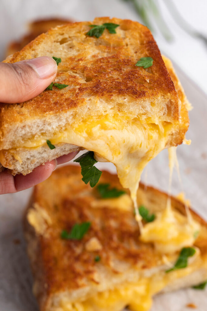 A hand holding up half of a sourdough grilled cheese with cheddar and swiss oozing out of the sandwich.